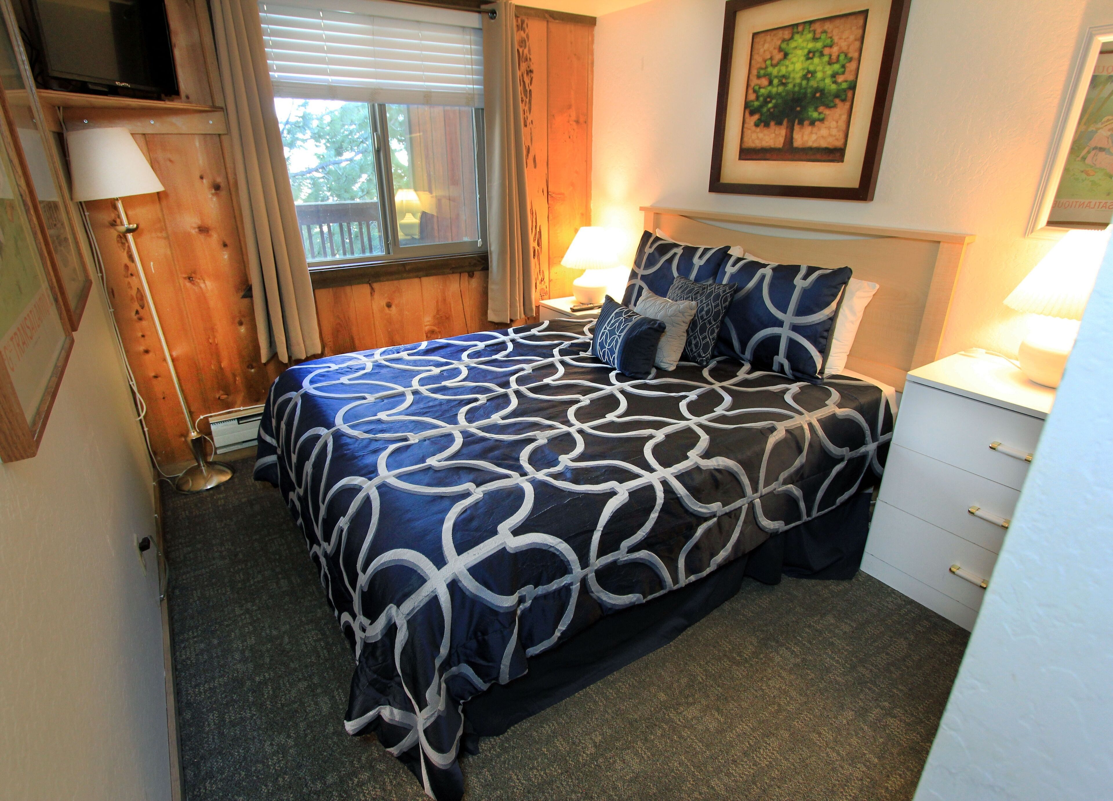 A bedroom with a bed, bedside lamps, a nightstand, a framed picture, and a window with blinds.