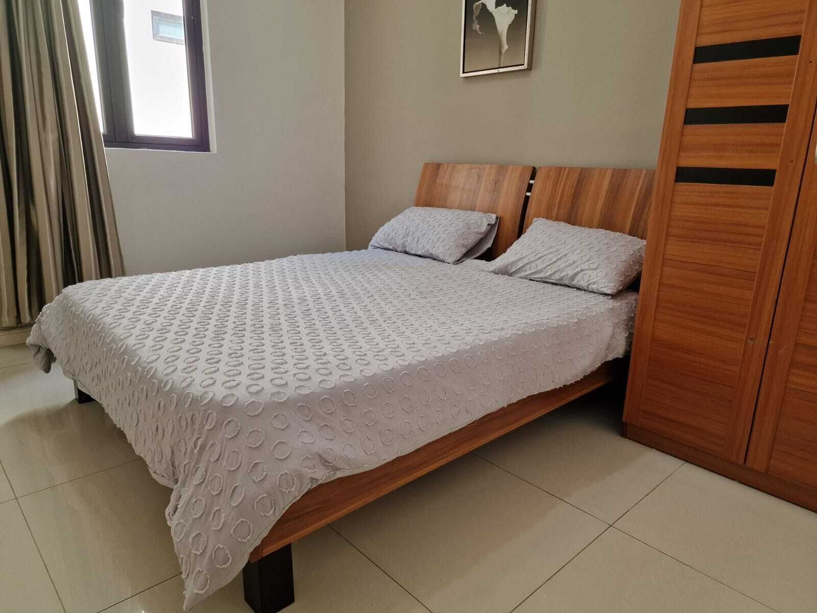 A neatly made bed with a patterned bedspread, wooden headboard, and a wooden wardrobe to the right.
