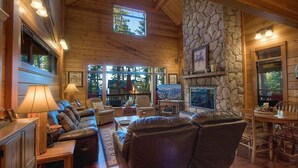 Interior acogedor de una cabaña de troncos con chimenea de piedra, asientos de cuero y ventanas amplias con vistas al exterior.