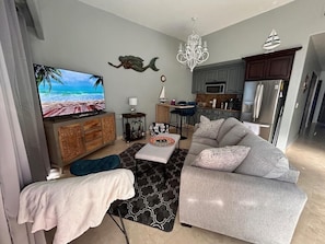 Ein modernes Wohnzimmer mit grauem Sofa, einem Flachbildfernseher, einer Holzkommode und einer Küchenzeile mit grauen Schränken und Edelstahlgeräten.