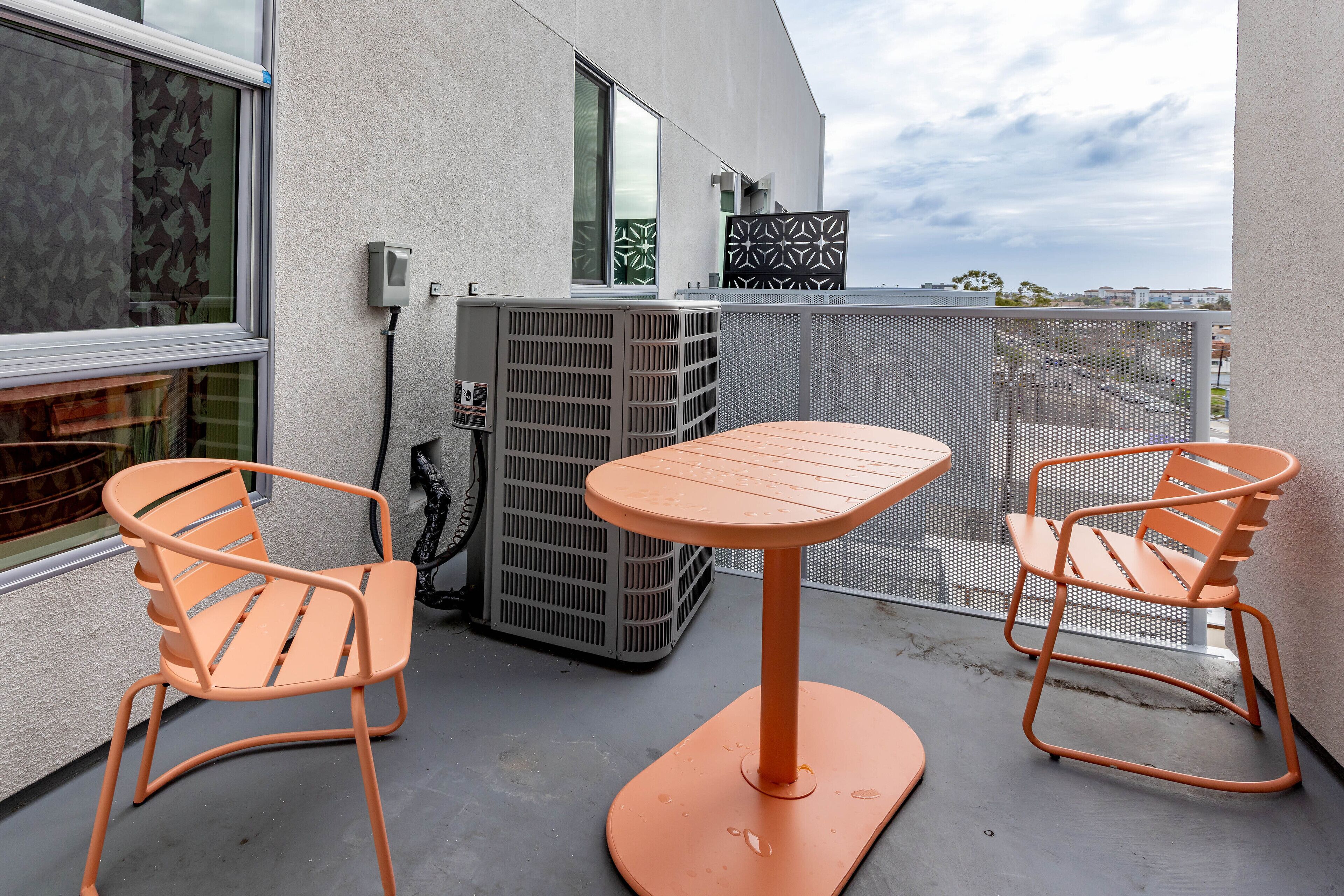 Ein Balkon mit zwei orangenen Stühlen, einem Tisch, einer grauen Klimaanlage und Blick auf die Stadtlandschaft.