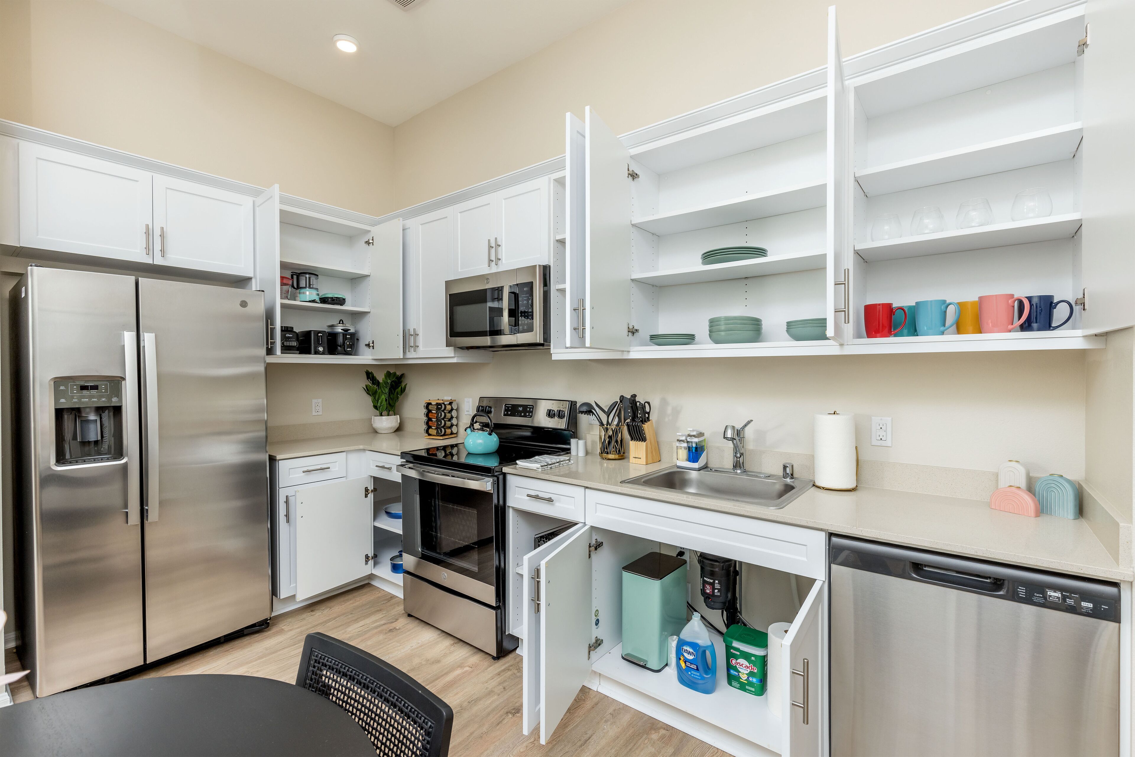 Una cocina moderna con armarios blancos, electrodomésticos de acero inoxidable y una variedad de tazas de colores en los estantes.