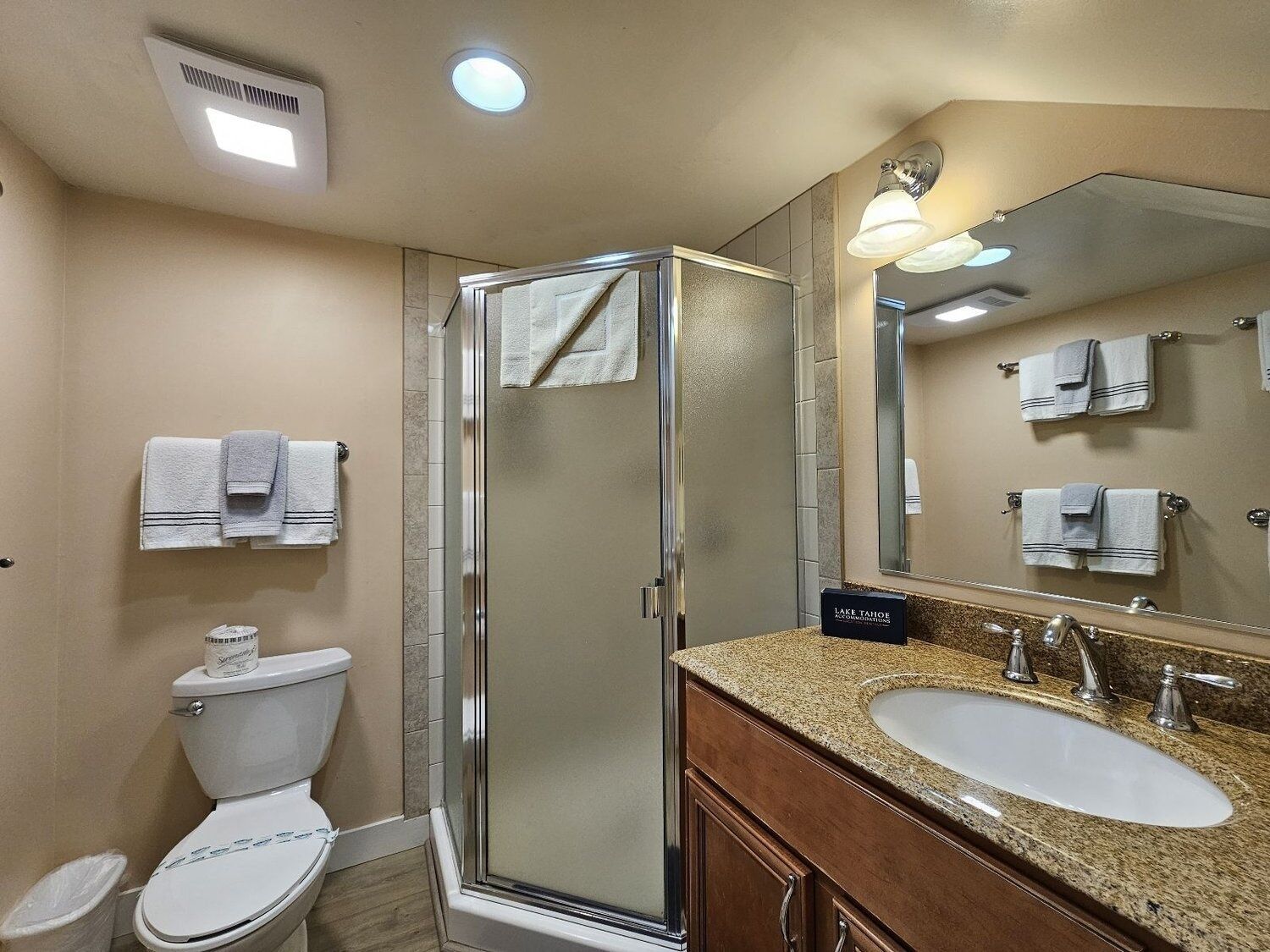 A hotel bathroom with a glass shower enclosure, a toilet, a sink with a granite countertop, and a mirror with towels hanging on the wall.