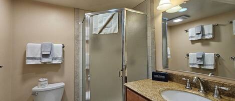 A hotel bathroom with a glass shower enclosure, a toilet, a sink with a granite countertop, and a mirror with towels hanging on the wall.