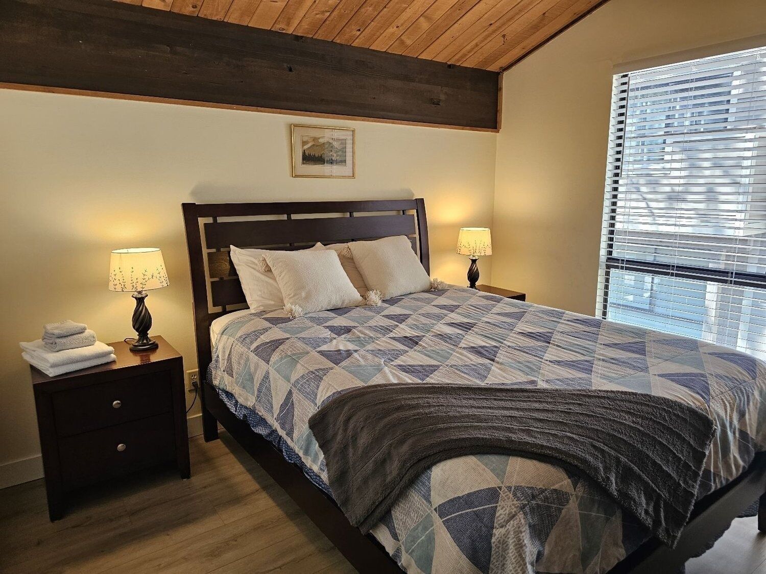 Une chambre avec un plafond en bois, un lit recouvert d’une courtepointe à motifs, deux lampes de chevet, une table de chevet avec une lampe et une fenêtre avec des stores.