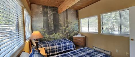 A bedroom with two beds, a nightstand, a lamp, and a mural of a forest on the wall.