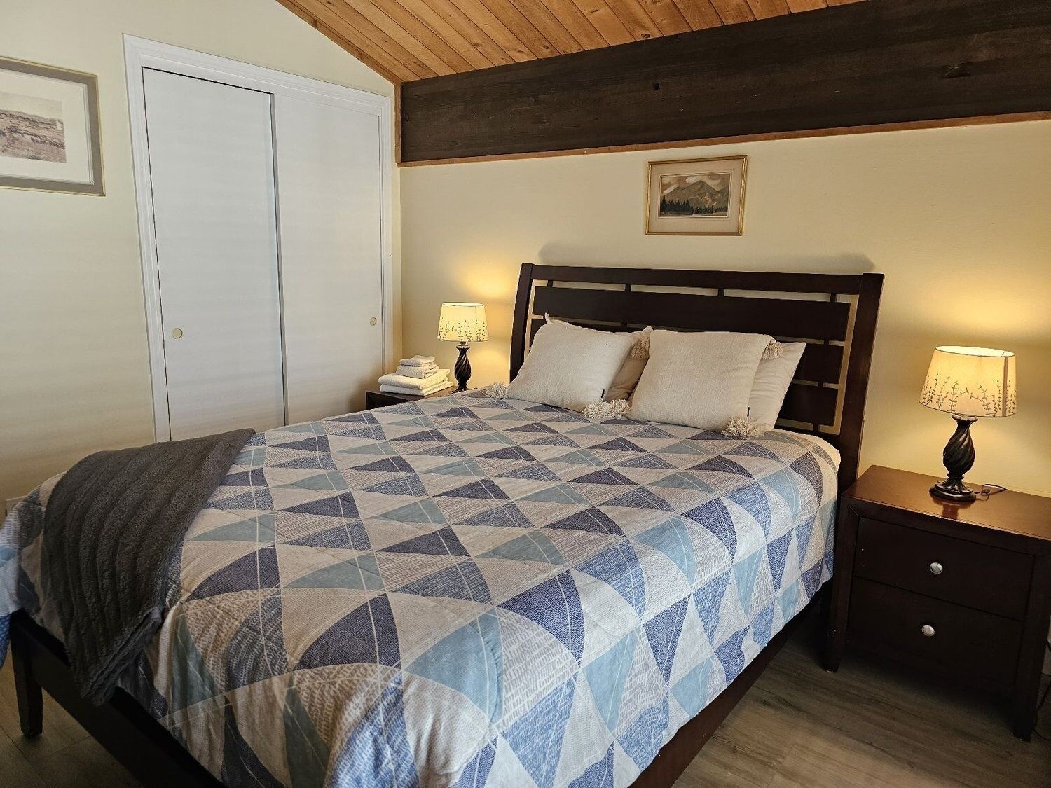 Un dormitorio con una cama, dos mesitas de noche, una mesita auxiliar, techo de madera y un cuadro enmarcado en la pared.