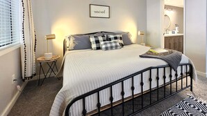 A bedroom with a bed, bedside lamps, a bedside table, a window with curtains, a framed picture on the wall, and a bathroom visible through an open door. - Taste Of Burgundy 1 Bedroom Condo by RedAwning (Incline Village)