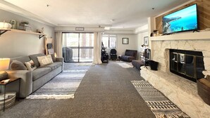 A living room with a sofa, a fireplace, a television, and a view through the glass doors. - Taste Of Burgundy 1 Bedroom Condo by RedAwning (Incline Village)