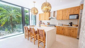 Una cocina moderna con armarios de madera, una isla central con taburetes altos y un amplio ventanal con vistas a un jardín tropical.