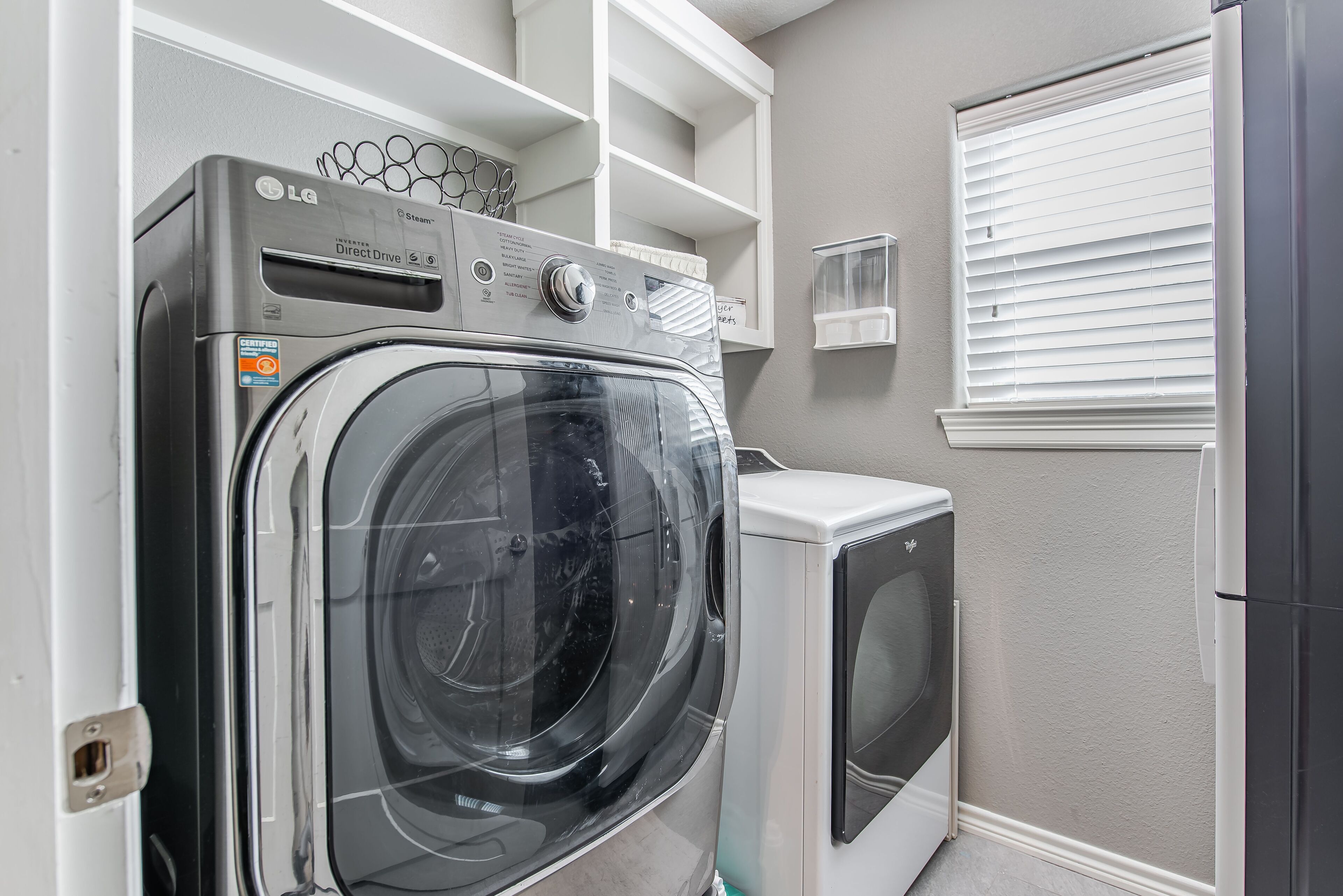 Une buanderie équipée d’un lave-linge et d’un sèche-linge à chargement frontal, d’étagères intégrées et d’une fenêtre avec des stores.