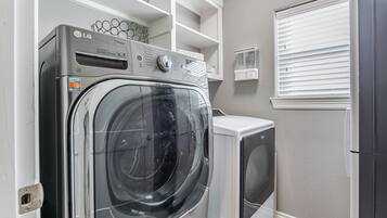 Une buanderie équipée d’un lave-linge et d’un sèche-linge à chargement frontal, d’étagères intégrées et d’une fenêtre avec des stores.