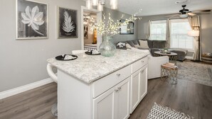Een moderne keuken met een marmeren aanrechtblad, witte kasten en een centraal eiland. Op het eiland staat een vaas met bloemen en erboven een plafondventilator.