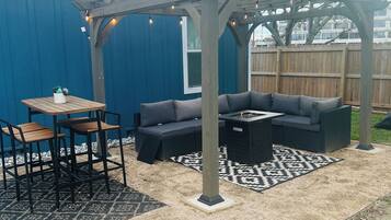 A covered patio area with a wooden table and chairs, a black outdoor sofa, and a fire pit.