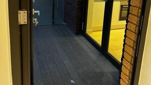 Ein schmaler Außenkorridor mit Holzboden, einer Glastür zu einem hellen Innenraum und einer Ziegelwand.