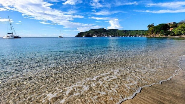 Une plage aux eaux claires et peu profondes, un voilier et une montagne qui se dessine à l’horizon.