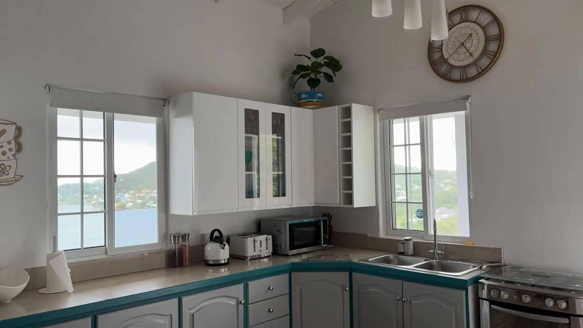 Eine Küche mit weißen Schränken, einer Mikrowelle, einem Toaster und einem Herd. Fenster bieten Blick auf eine Landschaft.