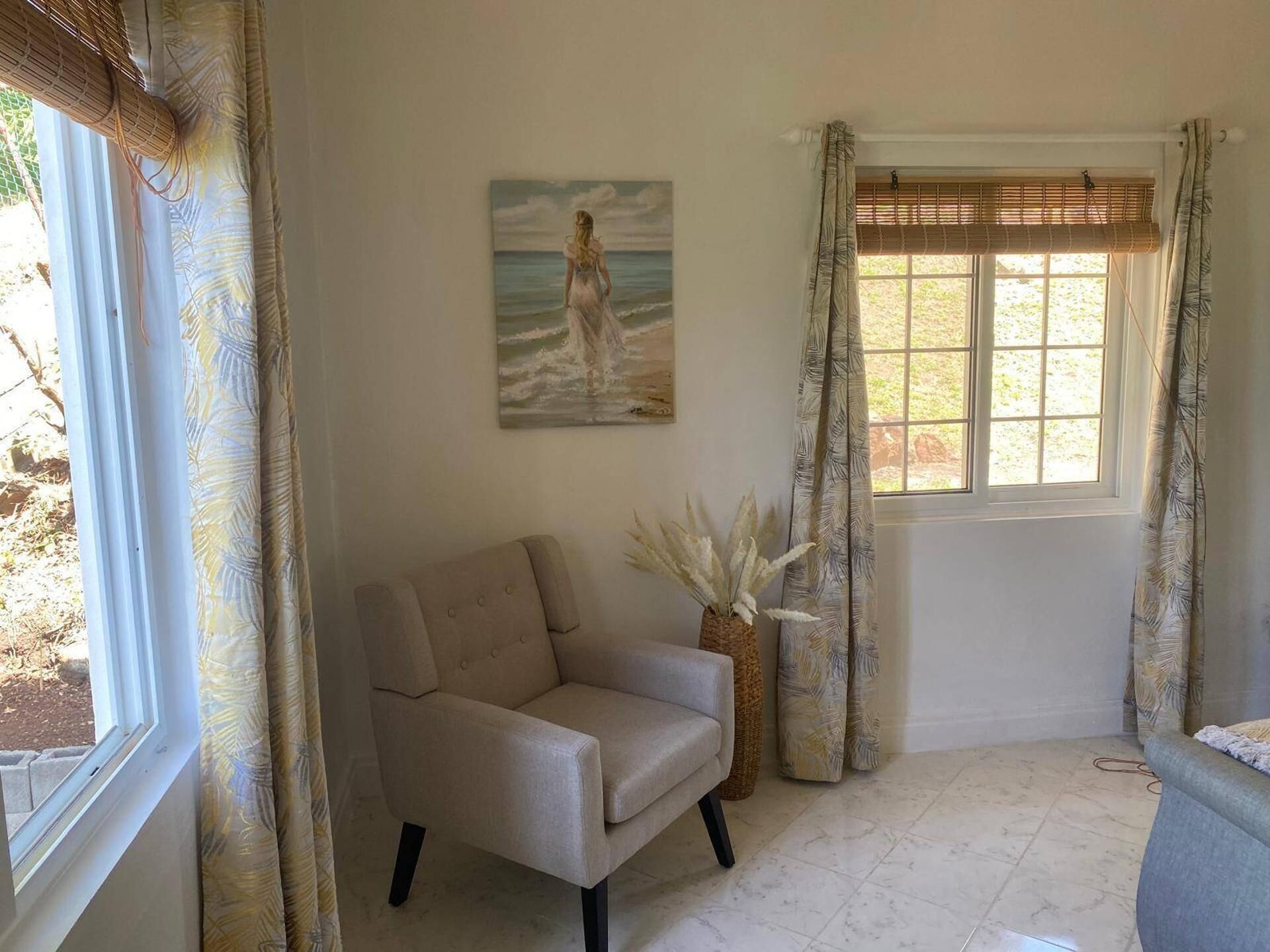 Ein Zimmer mit einem beigefarbenen Sessel, einem Bild einer Frau am Meer, einem Fenster mit Jalousien und einer Vase mit weißen Blumen.