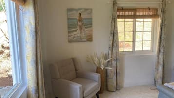 Ein Zimmer mit einem beigefarbenen Sessel, einem Bild einer Frau am Meer, einem Fenster mit Jalousien und einer Vase mit weißen Blumen.