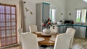 Un coin repas avec une table en bois, des chaises blanches, un vase de fleurs et une cuisine en arrière-plan.