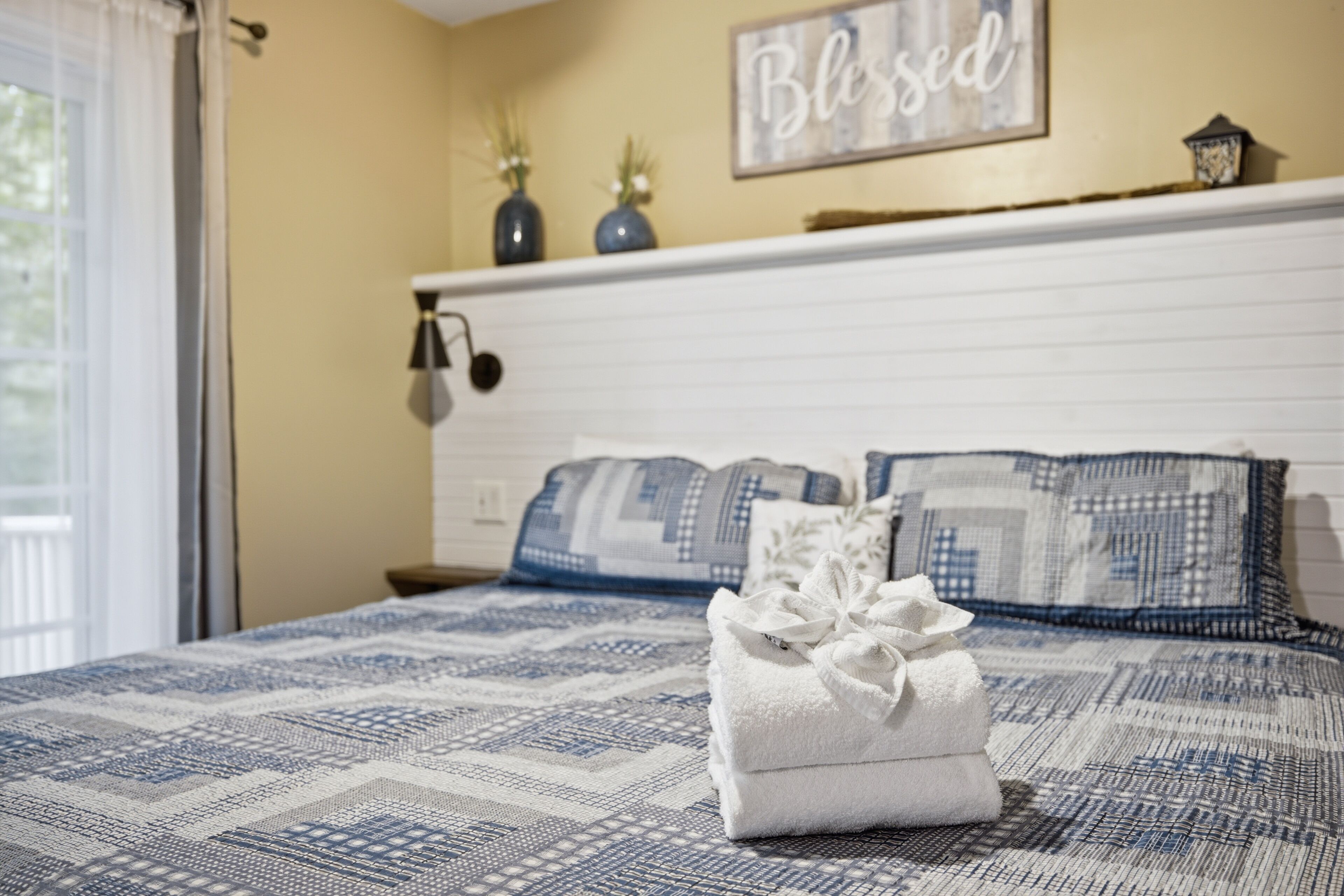 A neatly made bed with a patterned bedspread, a white towel with a bow, and a wall with a 'Blessed' sign.