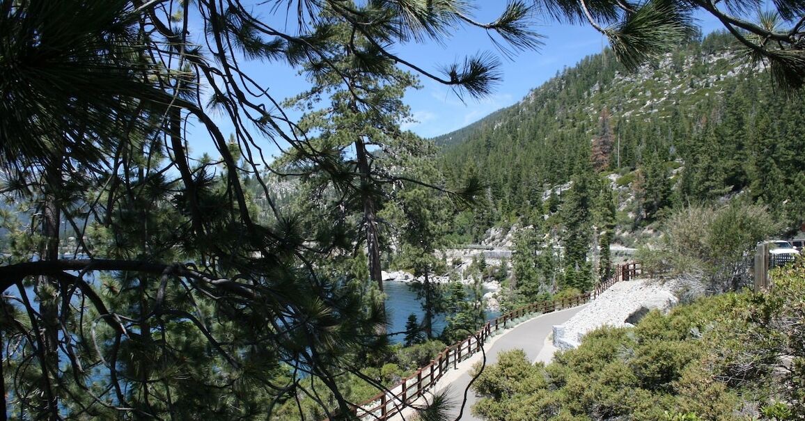 A scenic view of a lake surrounded by dense forest and mountains. A wooden pathway leads to a rocky area.