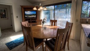 Un coin repas avec une table ronde en bois, six chaises et un lustre. Un téléviseur est fixé au mur, et il y a une grande fenêtre avec des rideaux.
