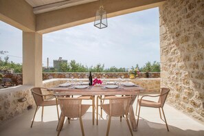 Un espacio de comedor al aire libre con techo, con un muro de piedra de fondo, una mesa preparada para cuatro personas y sillas de mimbre.