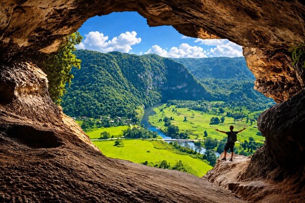 Una persona sobre una roca, con vista a un valle y un río que se ven a través de un arco natural.
