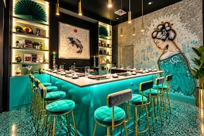 Un espace bar avec des tabourets de bar de couleur turquoise, un comptoir avec des verres et une fresque représentant une femme.