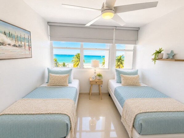 Une chambre avec deux lits, un ventilateur de plafond, une fenêtre donnant sur la plage et une petite table de chevet avec une lampe.
