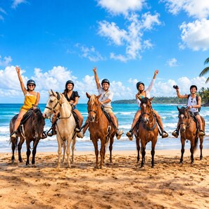 Un groupe de personnes à cheval sur une plage de sable, avec l’océan et un ciel dégagé en arrière-plan.