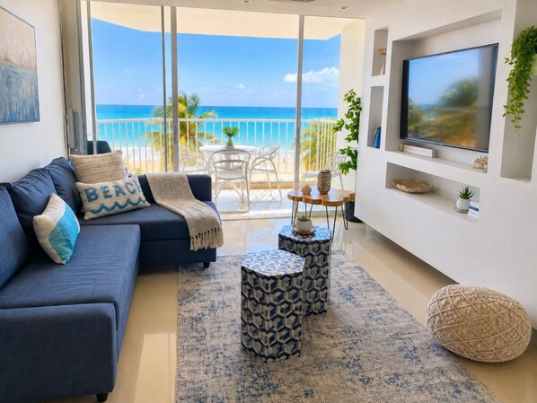 A living room with a sofa, coffee table, and TV, overlooking a balcony with a view of the ocean. - Beachfront 2b/2b King Bed Isla Verde Parking (Carolina)