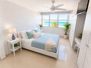 Een slaapkamer met een bed, nachtkastje, plafondventilator, raam met uitzicht op de oceaan en een kledingkast.