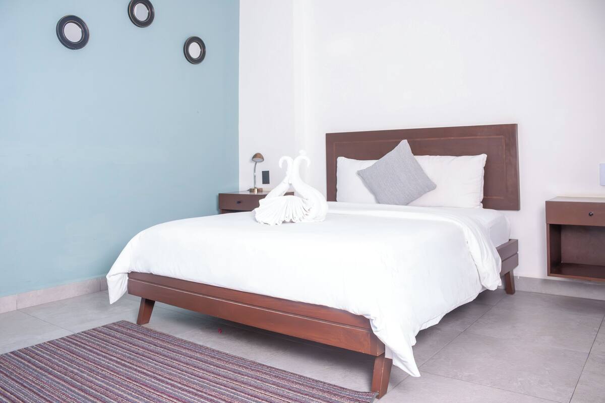 Un lit bien fait, avec une couette blanche et des oreillers, une tête de lit en bois, une table de chevet avec une lampe et un tapis à rayures sur un sol carrelé.