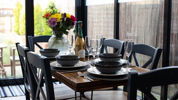 A dining table set with black bowls, plates, and wine glasses, a bottle of Prosecco, and a vase with colorful flowers. - Trinity Cottage, Stratford-upon-Avon - sleeps 5 guests in 3 bedrooms (Stratford-upon-Avon)