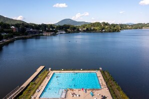 An aerial view of a swimming pool surrounded by a dock, with a lake and mountains in the background.