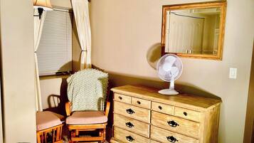 Une chambre avec une commode en bois, une chaise, un lit, une fenêtre avec des rideaux et un miroir au mur.