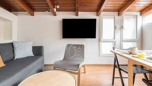 A modern living room with a grey sofa, a wooden dining table, and a grey armchair. A flat-screen TV is mounted on the wall.