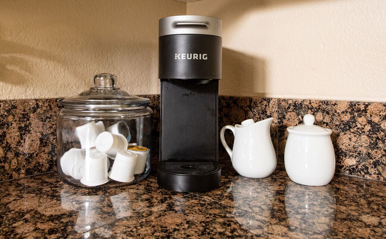 A coffee maker, a glass jar with cups, and two white pitchers on a granite countertop.