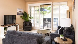 Un salon avec un canapé gris, une table basse, un téléviseur et une porte-fenêtre coulissante donnant sur une terrasse extérieure.