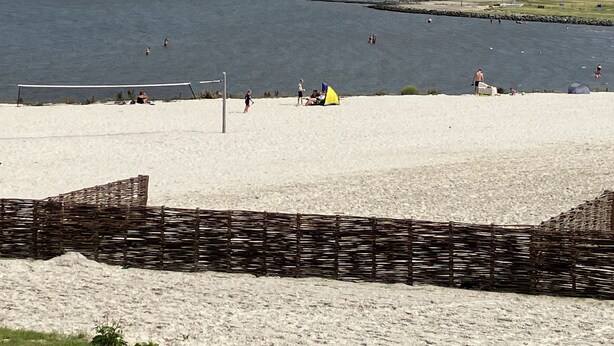 Una playa con una red de voleibol, una tienda de campaña amarilla y un faro al fondo.