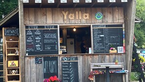 Un café al aire libre de madera llamado Yalla, con un cartel de menú, un letrero de bienvenida y un pequeño espacio de mesas.