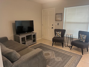 Un salon avec un téléviseur posé sur un meuble, deux canapés gris, un tapis à motifs et une porte ornée d’un tableau.