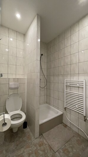 Bathroom features a white toilet, bathtub, and towel rack, set against a backdrop of large, square tiles.