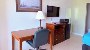 A hotel room with a wooden desk, a chair, a television, a dresser, and a small refrigerator.