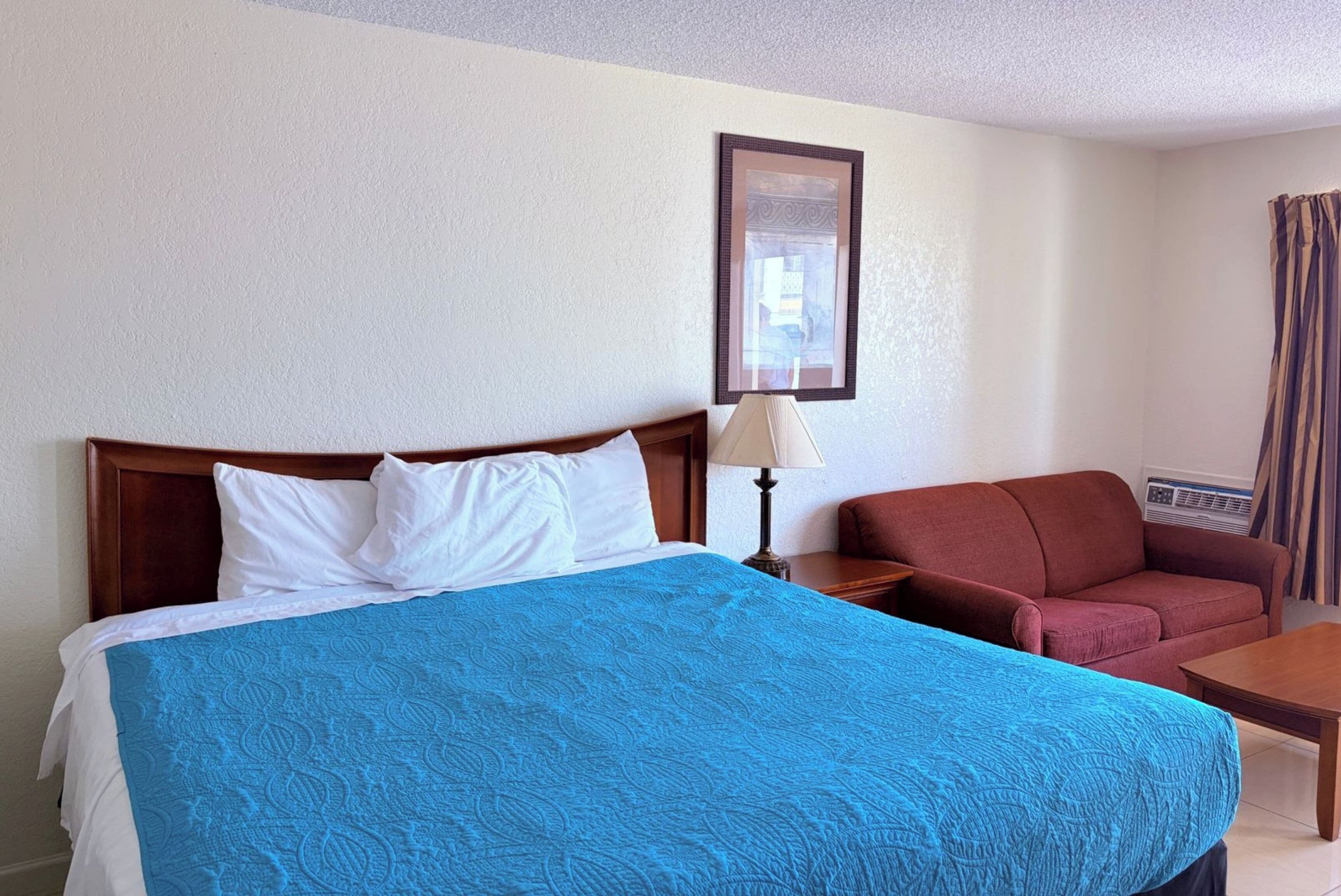 A hotel room with a bed, a red sofa, a wooden nightstand, a lamp, and a framed picture on the wall.