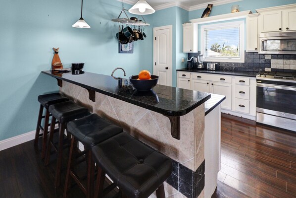A kitchen with a marble island, bar stools, a hanging pot rack, and a view of the outdoors through a window.