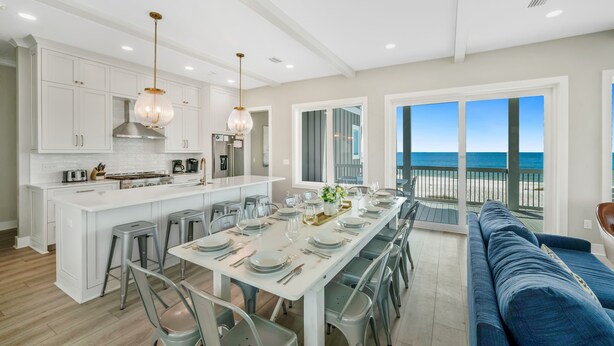 Une cuisine moderne avec des meubles blancs, un îlot central et un coin repas pouvant accueillir six personnes. Un balcon avec vue sur l’océan est visible.
