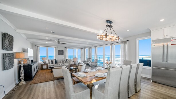 A spacious dining area with a wooden table set for six, surrounded by grey upholstered chairs. A modern chandelier hangs above, and the room features large windows offering a view of the ocean. - Ginger Snap: 5 BR Beachfront Home- Elevated Pool, Pet Friendly, Sleeps 20! (St. Joe Beach)